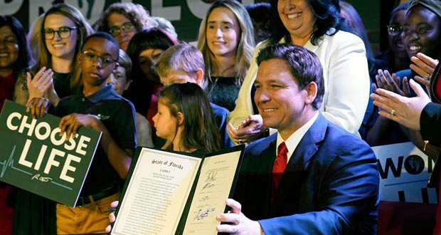 Ron DeSantis, sitting at a table, holds up a signed abortion ban law while surrounded by a mix of grown-ups and children