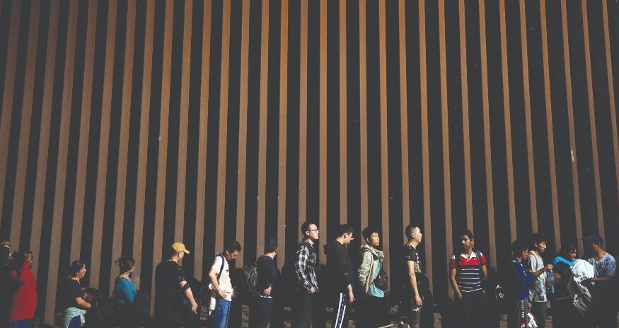 People line up against a border wall as they wait to apply for asylum after crossing the border from Mexico on July 11, 2023, near Yuma, Arizona. (AP photo: Gregory Bull)