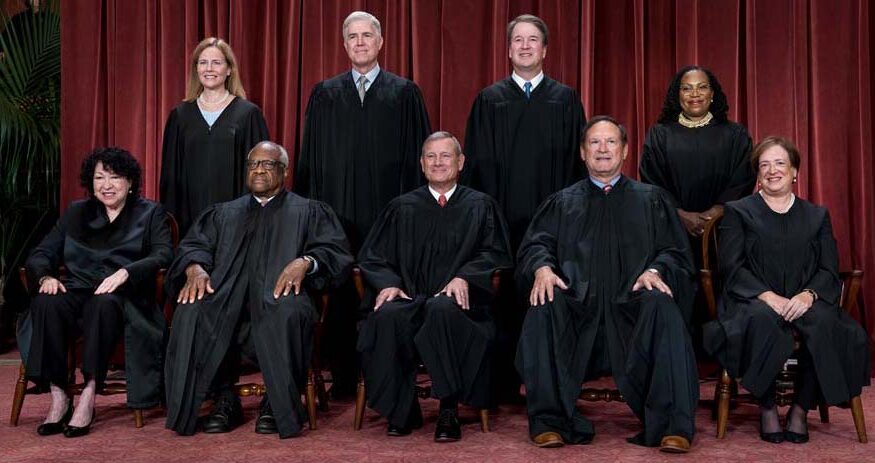 Current members of the U.S. Supreme Court stand and sit for a portrait in front of a red curtain