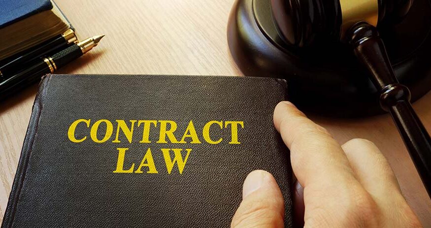 A book with a title of Contract Law resting on a desk with a person's hand near the right side of the book