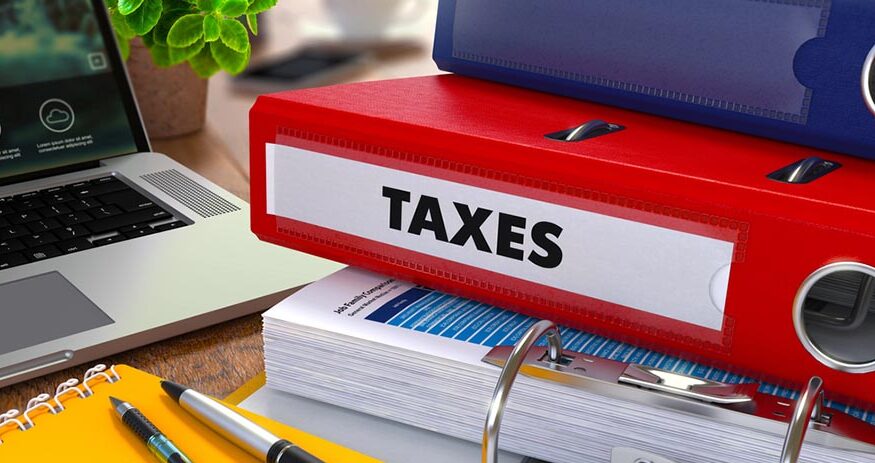 A stack of binders on an office desk with the word "TAXES" prominently displayed on a red binder edge