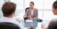 A conference table with an outside view behind, as a female lawyer facing the camera explains something to two people at the table facing her