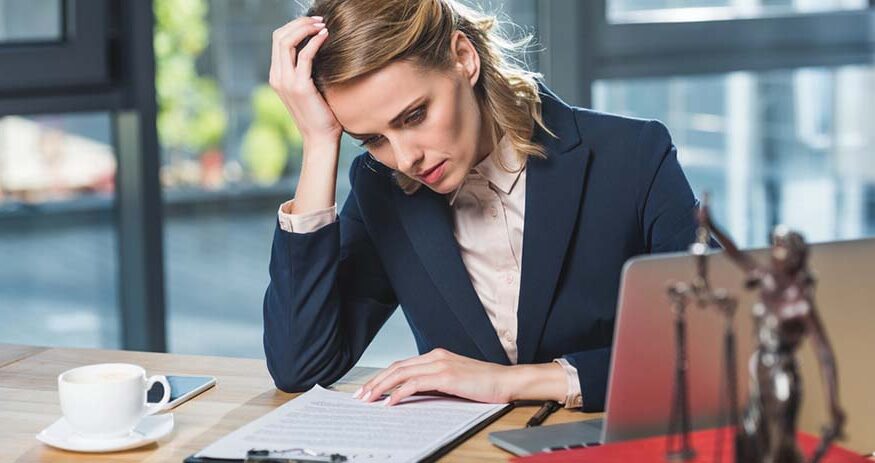 A woman in a business suit in front of a desk with her elbow on the desk and her head in her hand as she looks down at a paper with a tired look. A statue of a scales of justice is blurred in the foreground in the corner.